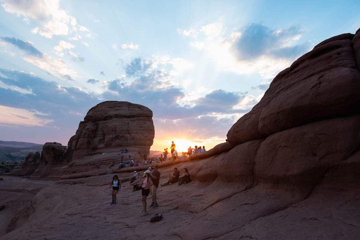 Hikers crossing the slickrock bowl toward Delicate Arch at sunset